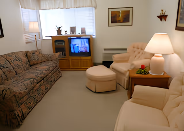 Living room with a patterned sofa, two upholstered armchairs and ottoman, a wooden TV cabinet, and table lamps.