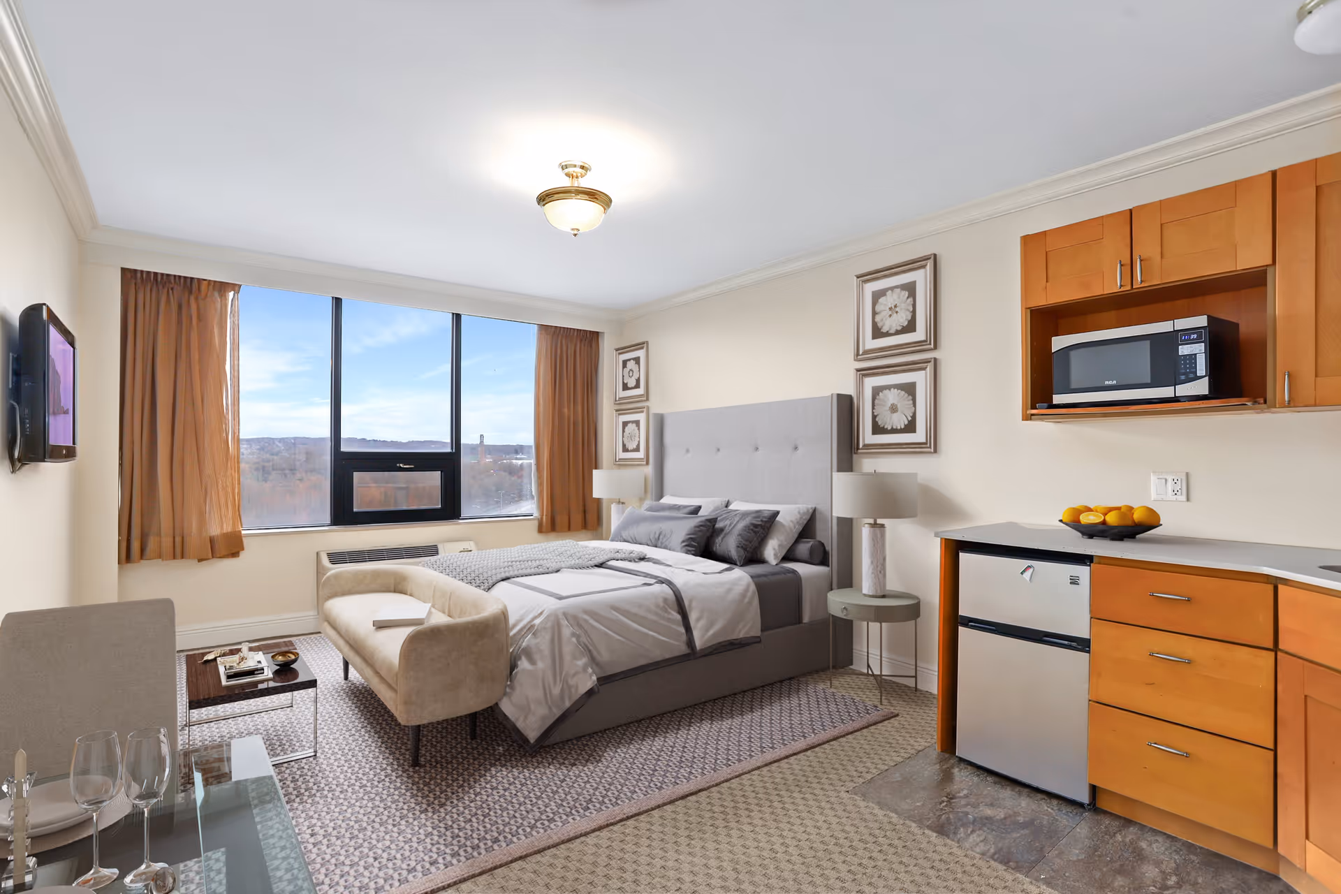 A bright and neatly arranged studio apartment featuring a bed with gray bedding and multiple pillows, a small beige loveseat at the foot of the bed, a side table with a lamp, and framed floral artwork on the wall. There is a large window with brown curtains letting in natural light and offering a view of trees and distant hills. The room also includes a kitchenette area with wooden cabinets, a microwave, a small refrigerator, and a bowl of oranges on the counter. A glass dining table with chairs and wine glasses is partially visible in the foreground.