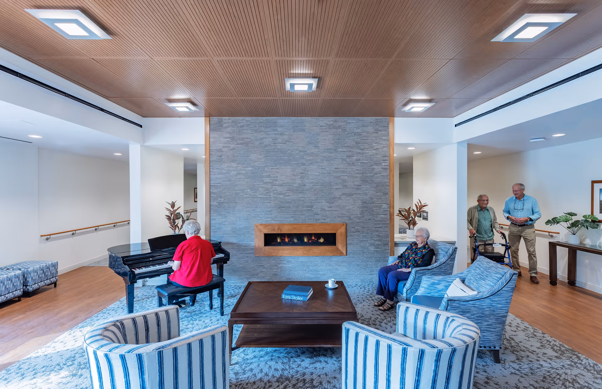 A senior living facility common area with a modern fireplace in the center of a gray tiled wall. An elderly woman in a red shirt is playing a black grand piano on the left side. Another elderly woman is seated in a blue armchair near the fireplace. Two elderly men, one using a walker, are standing and conversing on the right side. The room has a wooden ceiling with square light fixtures, wooden flooring, and several blue and white striped chairs arranged around a wooden coffee table with a book and a cup on it.