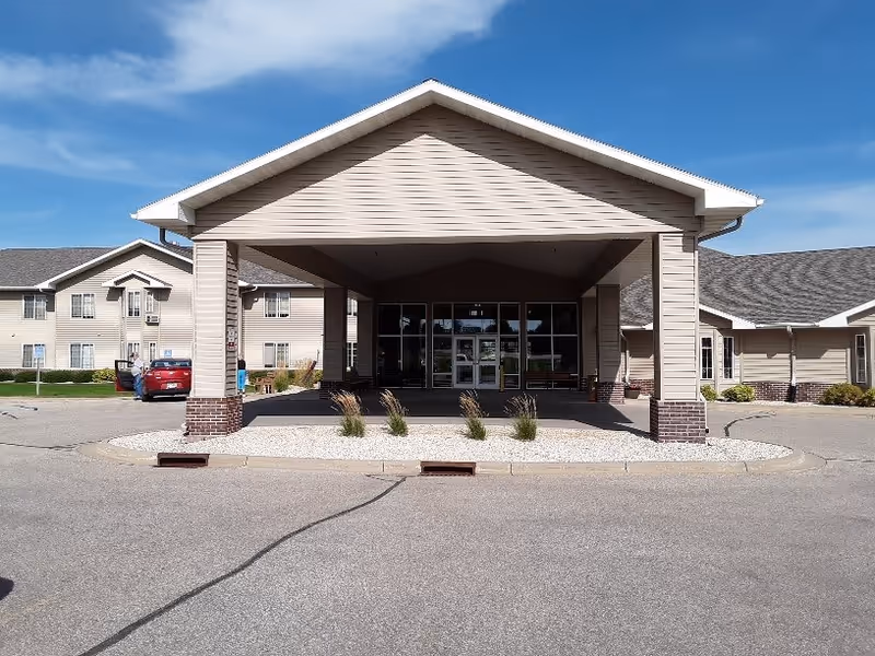 Front exterior view of Prairie Meadows Senior Living facility showing a covered entrance with pillars, a driveway, and beige siding buildings under a blue sky.