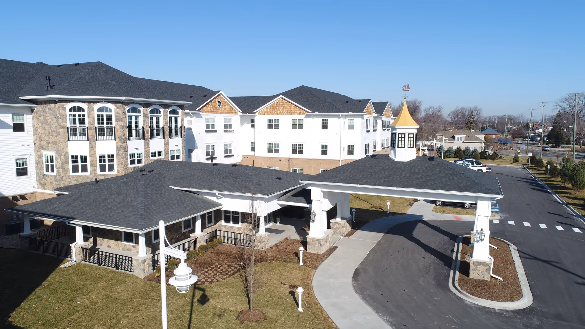Exterior view of the American House Lakeshore senior living facility showing a large multi-story building with white siding and stone accents, a covered entrance with a peaked roof and a weather vane on top, a driveway, parking spaces, and a clear blue sky.