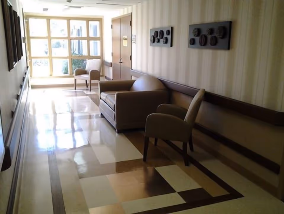 A well-lit hallway in a healthcare facility with a patterned floor, beige walls, and a large window at the end letting in natural light. The hallway features a brown loveseat and two beige armchairs along the right side, with decorative wall art above the seating.