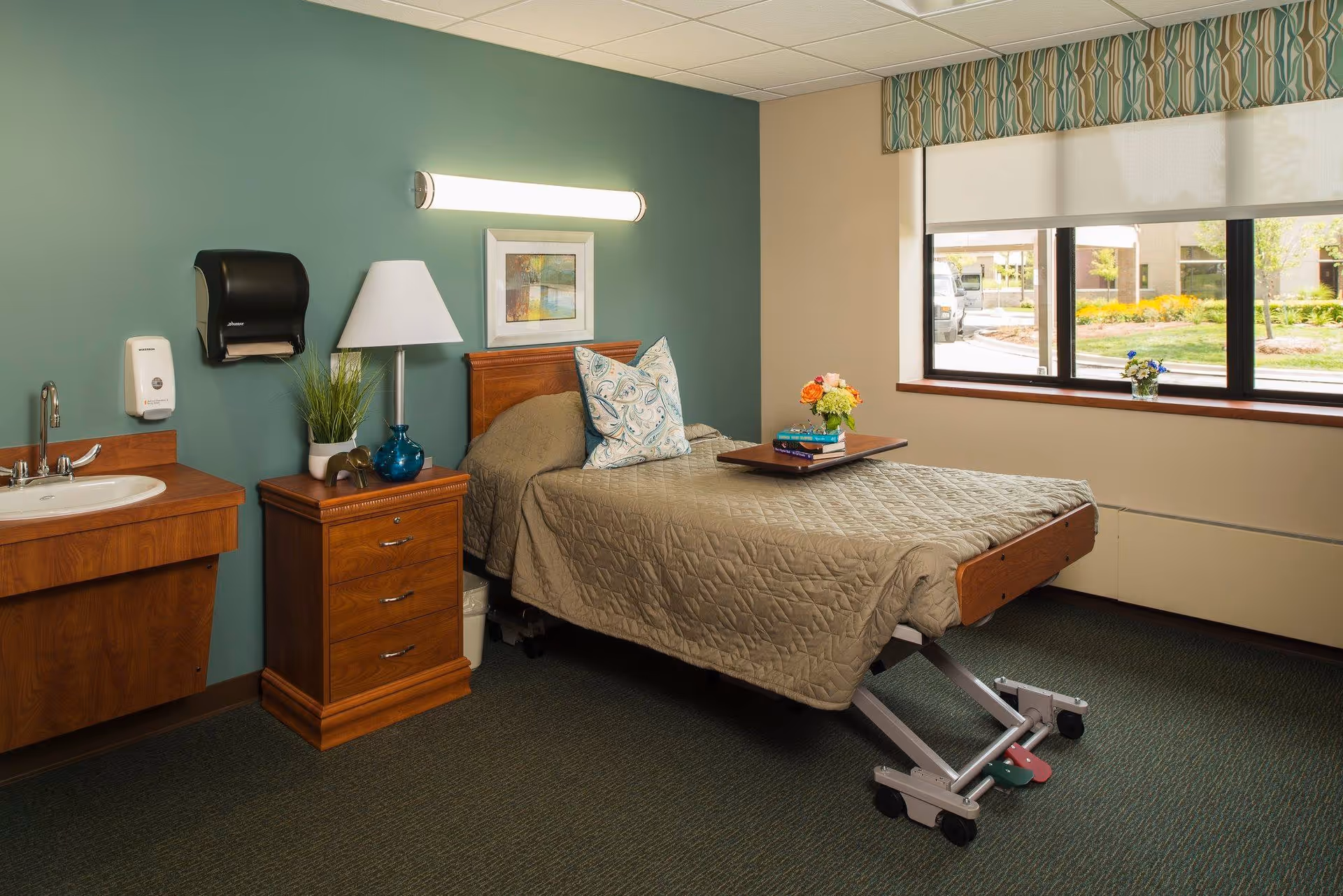 A tidy senior care bedroom with an adjustable bed, bedside table and lamp, a sink, and a window overlooking landscaped grounds.