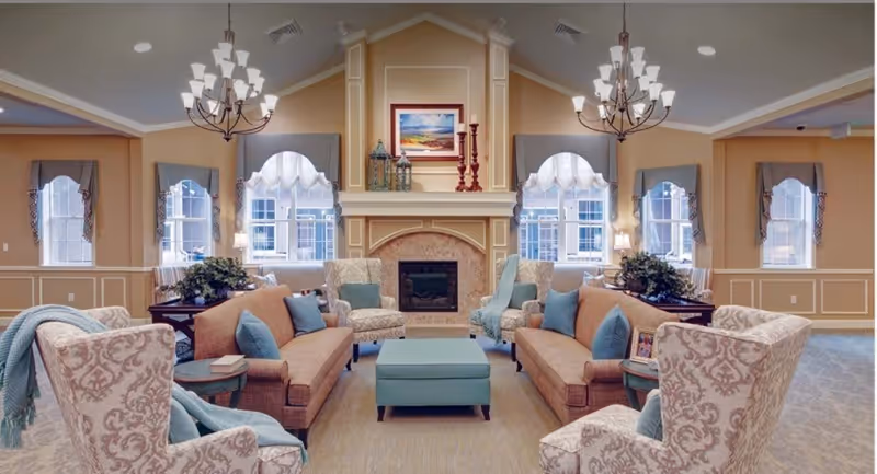 A spacious and elegantly decorated living room with two brown sofas facing each other, four patterned armchairs, a central blue ottoman, and a fireplace with decorative items and a framed painting above it. The room has large windows with white curtains, beige walls with white trim, and two chandeliers hanging from the ceiling.