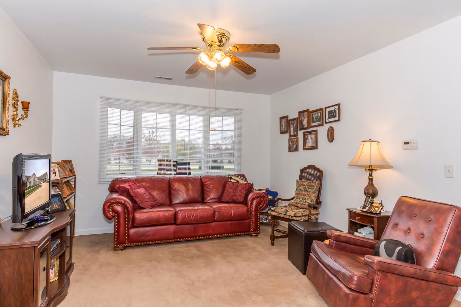 A cozy living room with a red leather sofa and matching armchair, a wooden side table with a lamp and telephone, a floral cushioned chair, a TV on a wooden stand, and framed photos on the wall. A ceiling fan with lights is mounted on the white ceiling, and a large window with blinds lets in natural light.