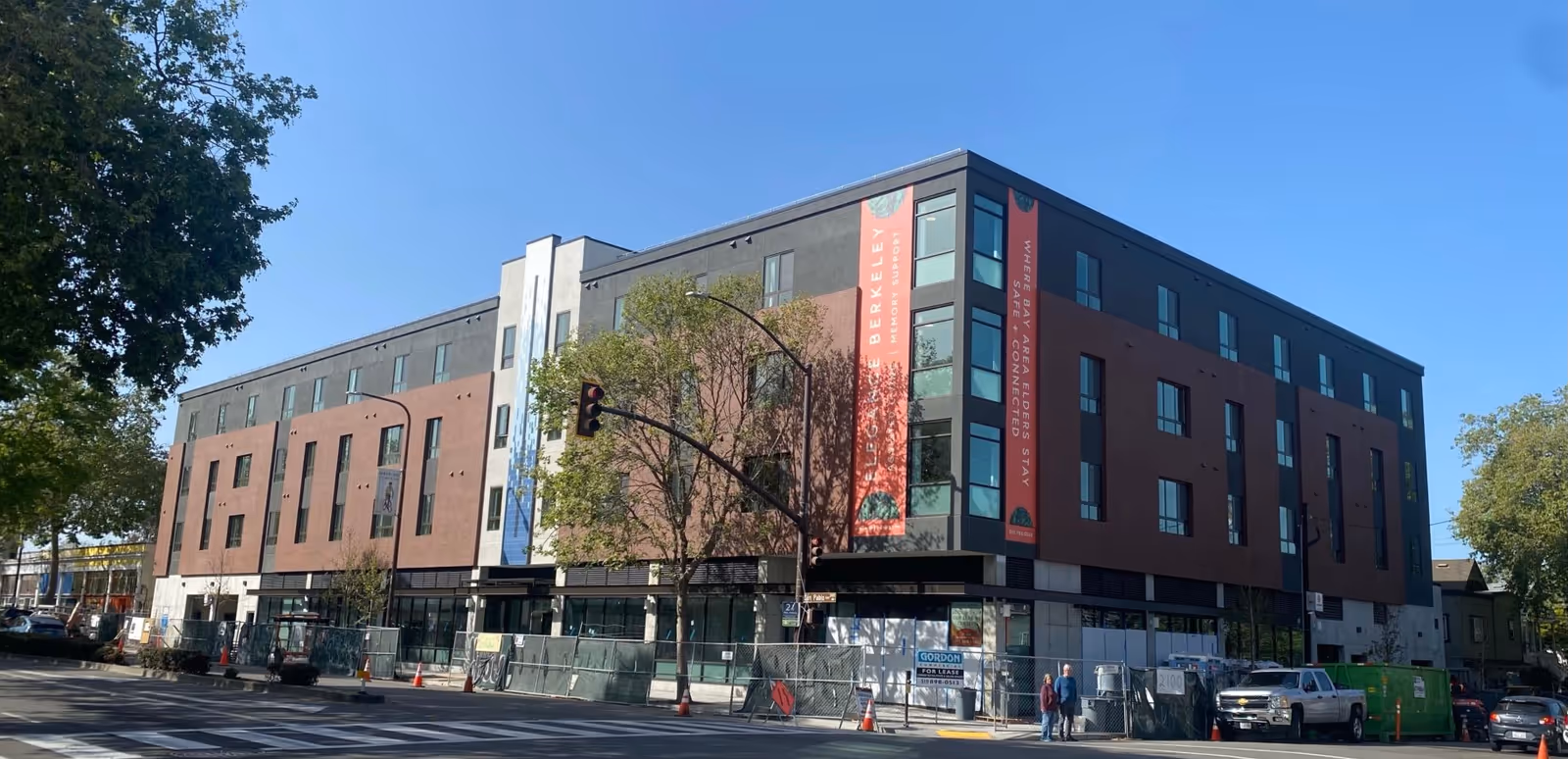 Four-story modern building on a street corner with large vertical banners and construction fencing around the ground level.