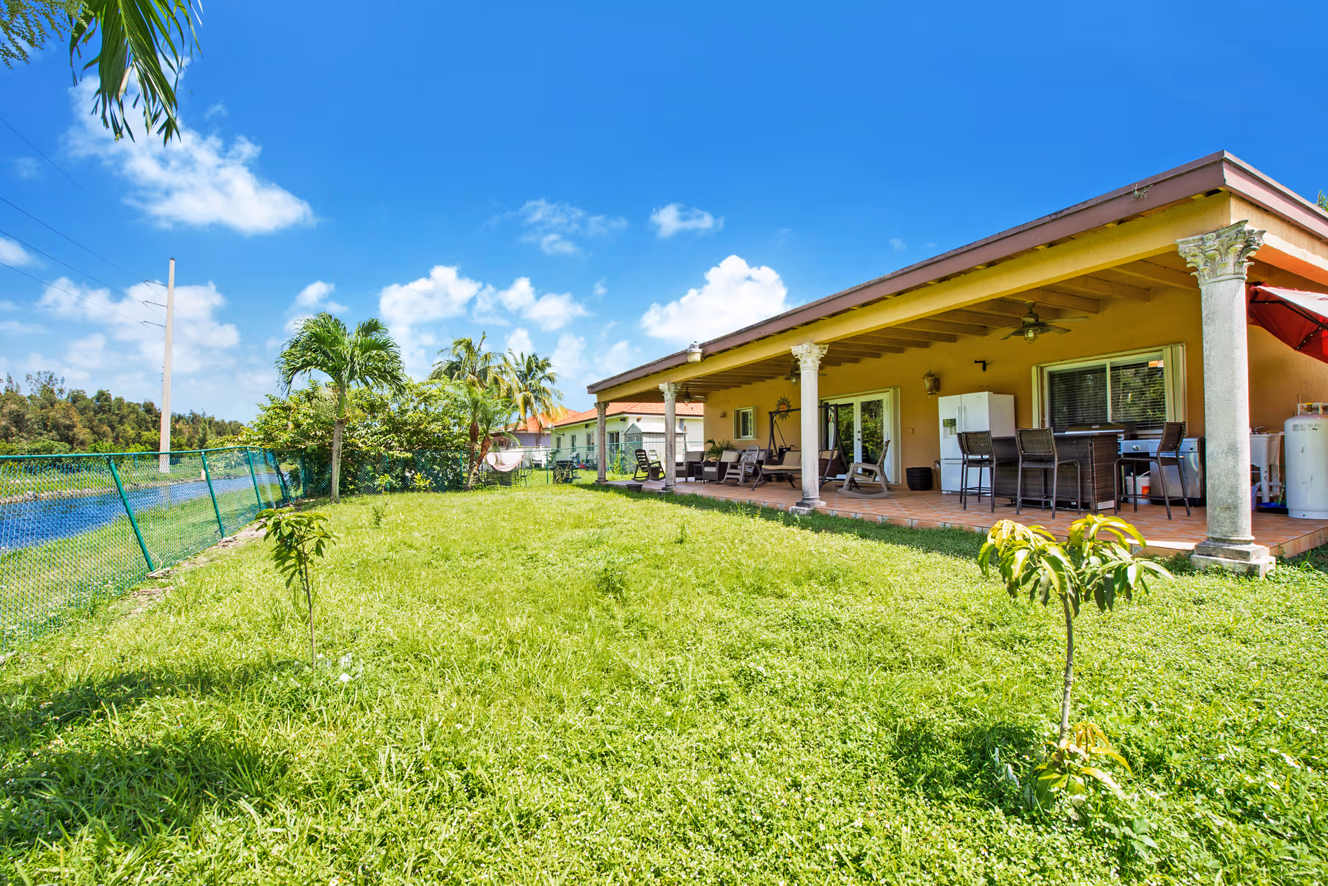 Grassy backyard with small trees and a covered patio attached to a single-story building under a blue sky.