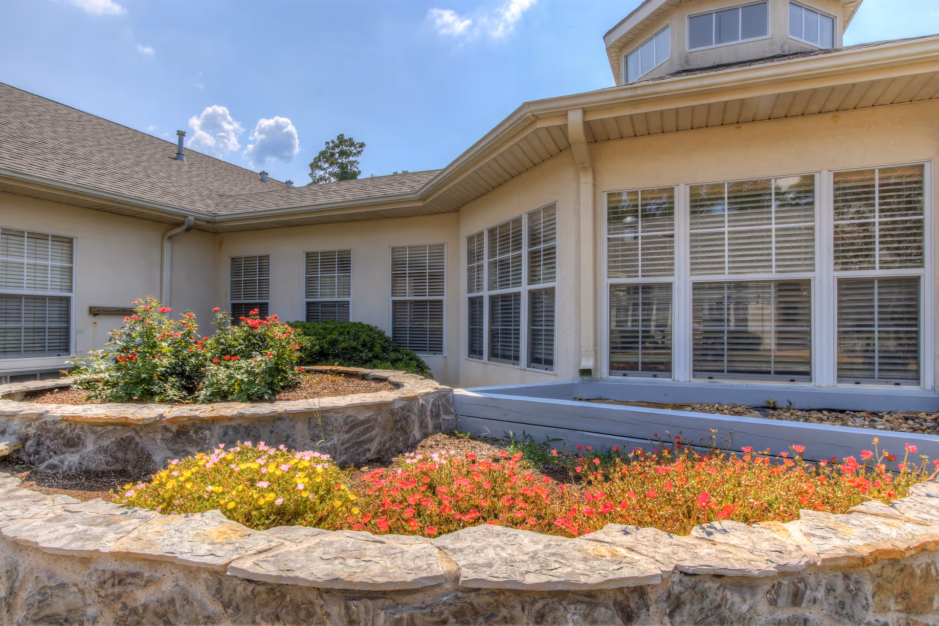 Exterior view of a senior living facility showing a stone-bordered flower bed with colorful flowers in front of a beige building with multiple windows and a clear blue sky above.