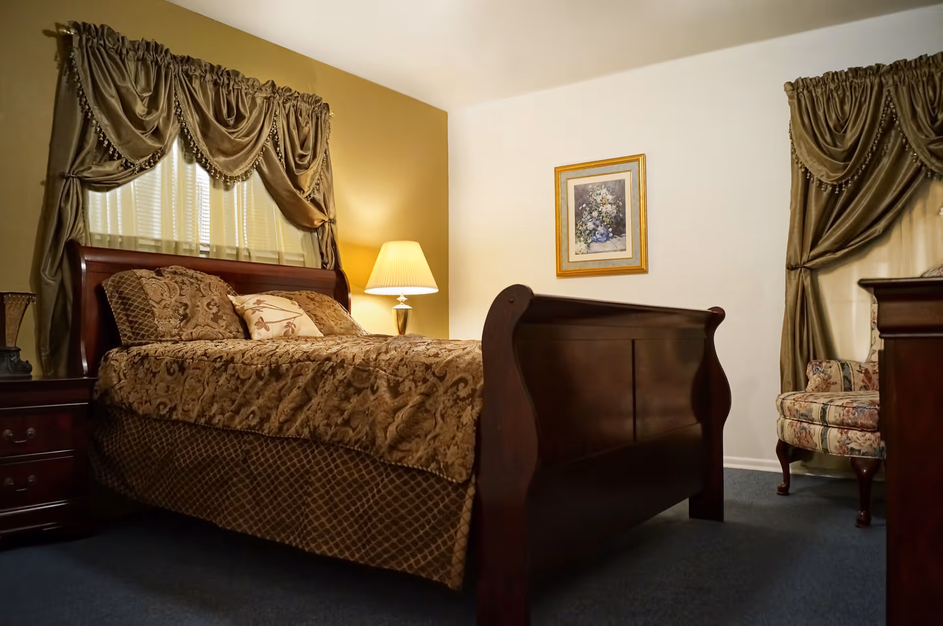 A bedroom with a wooden sleigh bed covered in a brown patterned bedspread and matching pillows. There are two windows with ornate brown curtains, a nightstand with a lamp, a framed floral painting on the wall, and a floral upholstered armchair in the corner.