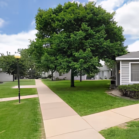 A well-maintained outdoor area with green grass, a large leafy tree, and a paved walkway leading through the space. Part of a building with a screened porch is visible on the right side under a partly cloudy sky.
