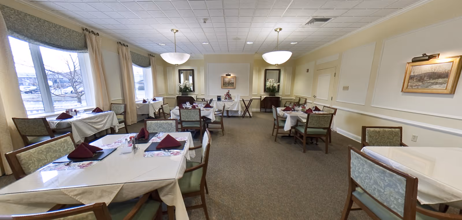 A dining room with several tables covered in white tablecloths and set with burgundy folded napkins, placemats, and glassware. The room has large windows with curtains, beige walls with framed artwork, and two hanging light fixtures from the ceiling. Chairs with patterned upholstery surround the tables.