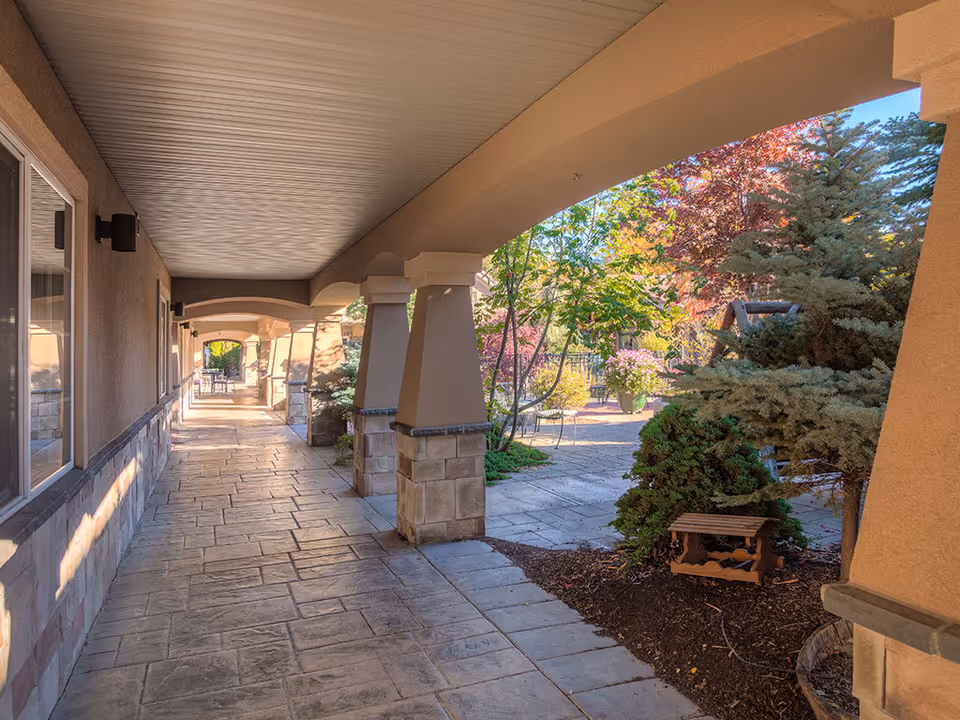 Covered outdoor walkway with stone columns and a tiled path opening onto a landscaped courtyard with trees and seating.