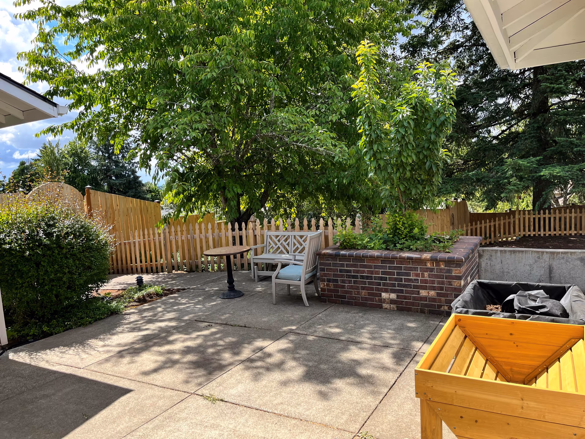 Outdoor patio area with concrete flooring, a wooden bench with cushions, a small round table, a raised brick planter with green plants, a wooden planter box, and a wooden picket fence surrounding the space. Trees and bushes provide shade and greenery under a partly cloudy sky.