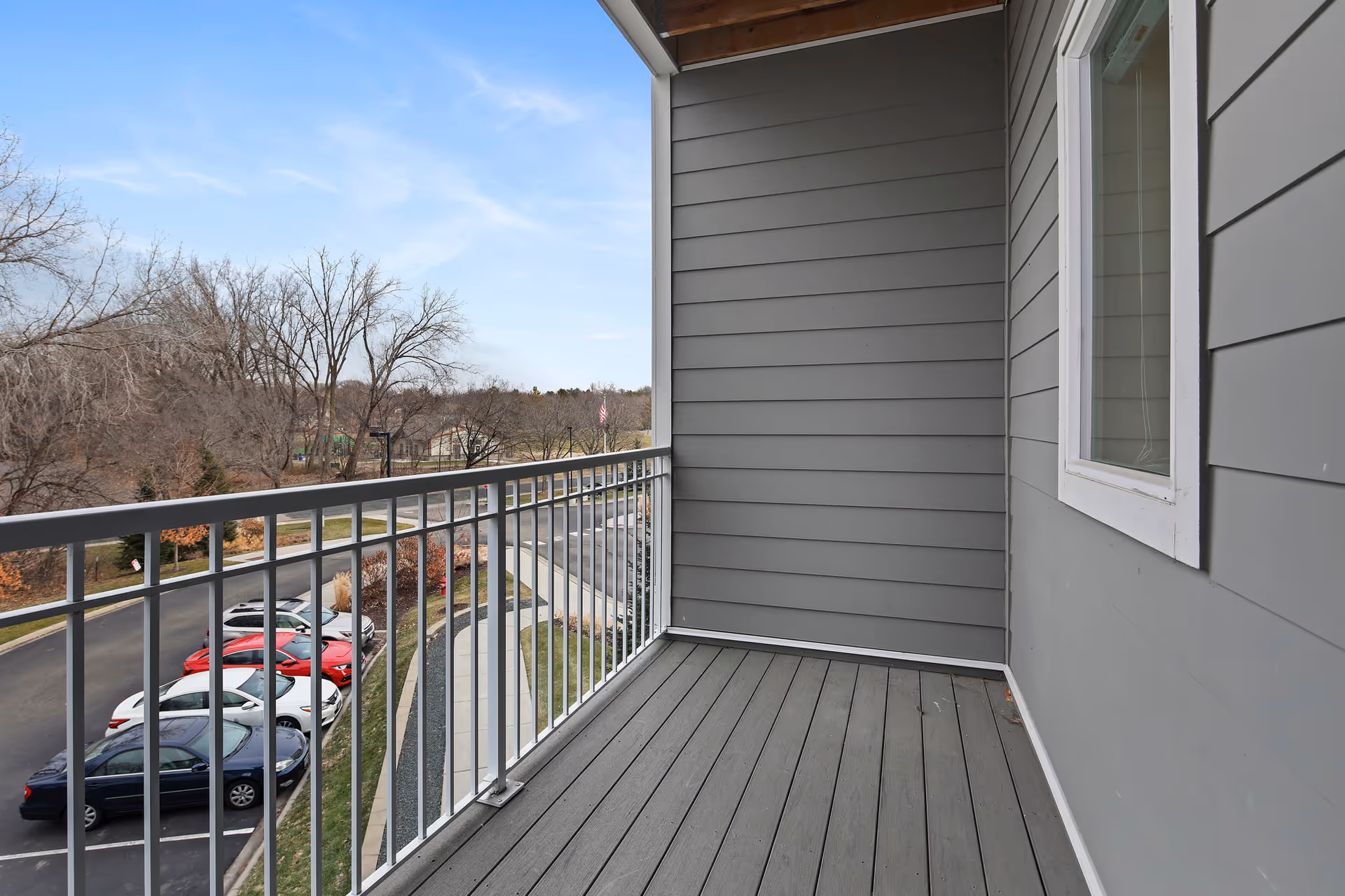 View from a balcony with gray wooden flooring and gray siding walls, overlooking a parking lot with several parked cars and leafless trees in the background under a clear blue sky.