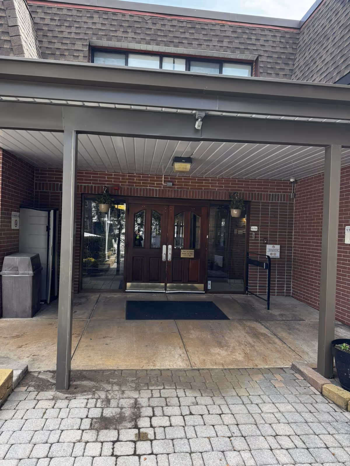 Entrance to Towson Rehabilitation and Healthcare Center featuring double wooden doors with glass panels, a covered walkway supported by metal posts, brick walls, and a paved stone pathway leading up to the entrance.