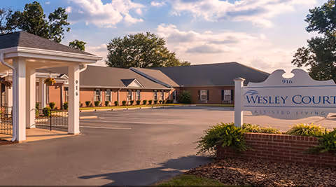Exterior view of Wesley Court Assisted Living Community showing a single-story brick building with a covered entrance and a large sign displaying the facility's name. The scene is set under a partly cloudy sky with trees in the background.
