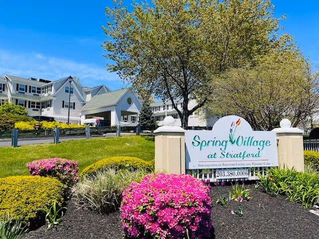 Exterior view of Spring Village at Stratford senior living facility with a large sign in the foreground surrounded by colorful flowers and landscaped bushes. The building is visible in the background under a clear blue sky.