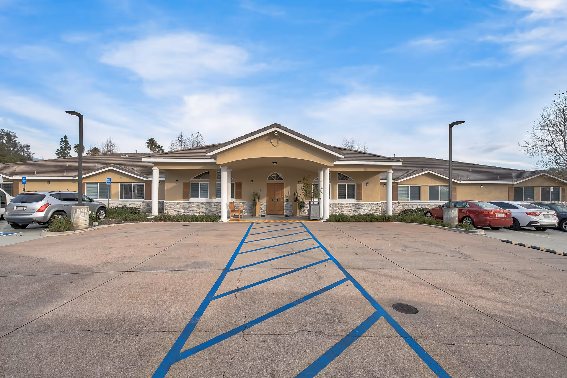 Front exterior view of a single-story assisted living facility building with a covered entrance supported by white columns. There are several parked cars on either side of the driveway, and the sky is partly cloudy.