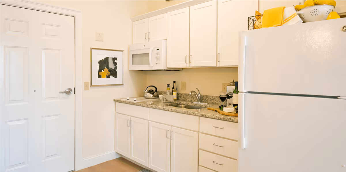 A small kitchen area with white cabinets, a granite countertop, a white refrigerator, and a white microwave mounted above the counter. On the counter are a kettle, bottles, a cutting board with cheese, and two glasses of wine. A framed abstract painting hangs on the wall next to a white door.