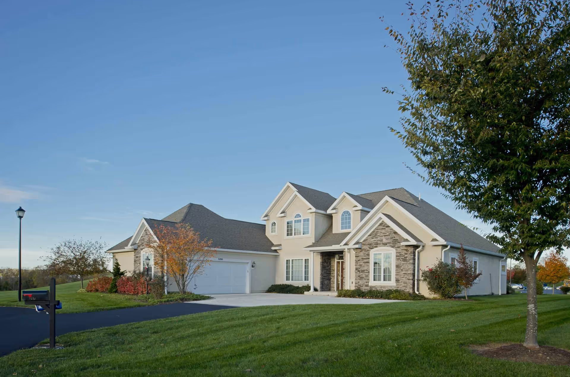 A large, modern two-story house with beige walls and stone accents on the front facade. The house has multiple windows, a double garage, and a well-maintained green lawn with trees and shrubs. The sky is clear and blue.