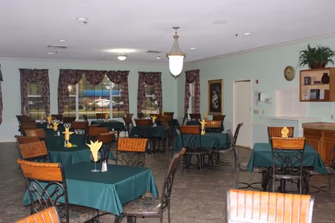 Dining room with multiple tables covered in green tablecloths, each set with yellow folded napkins. The room has several windows with floral curtains, a light fixture hanging from the ceiling, and wooden chairs with metal frames. There is a sideboard with shelves and decorative items on the right wall.