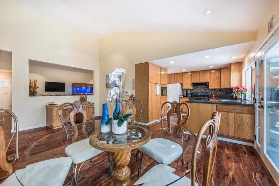 Interior view of a dining area and kitchen in an assisted living facility. The dining area features a round glass-top table with ornate wooden chairs that have light-colored cushions. The kitchen has wooden cabinets, a white refrigerator, a black stove, and a black countertop. There are decorative vases with flowers on the dining table and a window letting in natural light near the kitchen. The floor is wooden, and there is a wall-mounted TV and an aquarium visible in the background.