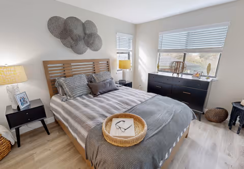 Well-lit bedroom with a wooden bed, bedside tables and lamps, a dresser under windows, and decorative wall art.