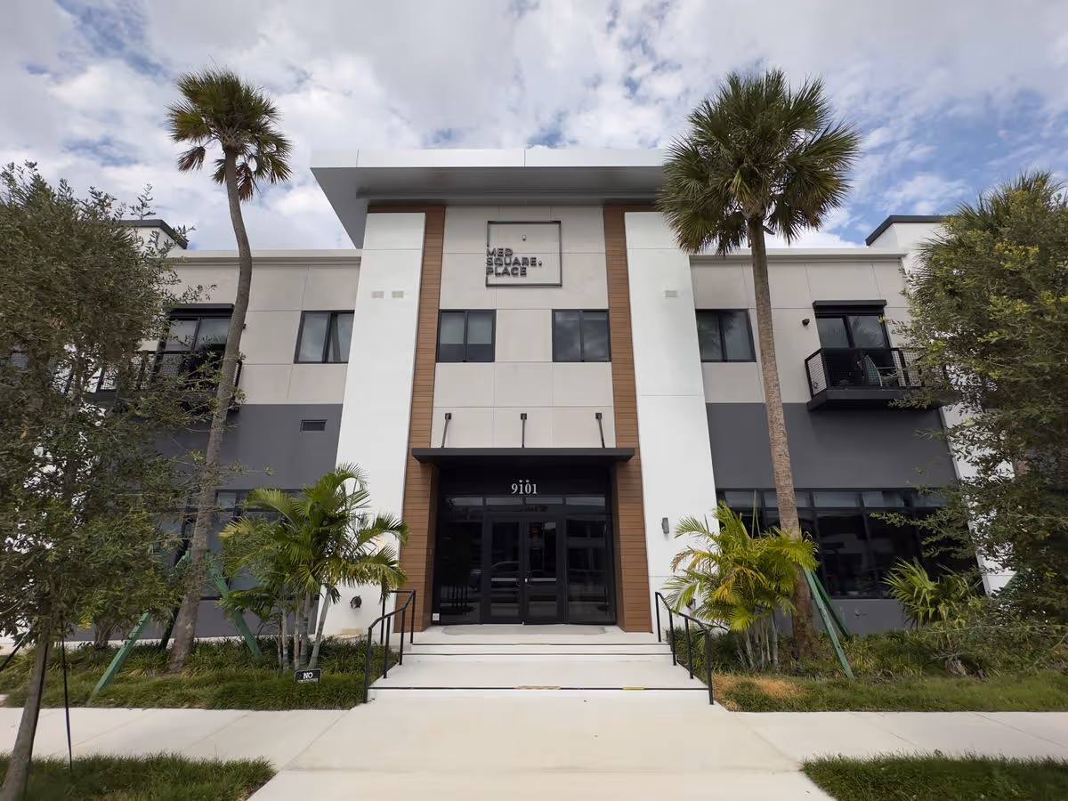 Front exterior of a modern two-story residential building with palm trees flanking steps up to the main glass entrance.