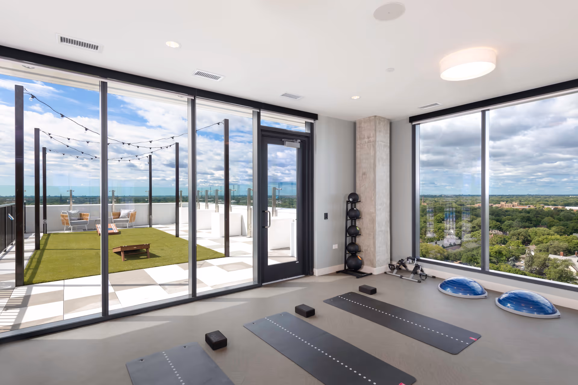 A bright fitness room with large floor-to-ceiling windows offering a panoramic view of a green landscape and cloudy sky. The room contains three black yoga mats with blocks, two blue balance trainers, a rack holding medicine balls, and a small exercise machine. Outside the glass door is a rooftop patio with string lights, artificial grass, and seating areas.