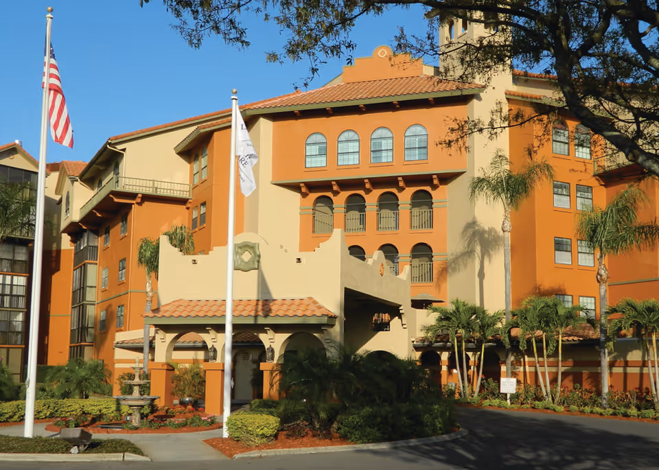 Exterior view of Lake Seminole Square, a multi-story senior living facility with a terracotta roof, beige and orange walls, arched windows and entrance, surrounded by palm trees and landscaping, with two flagpoles displaying the American flag and a white flag.