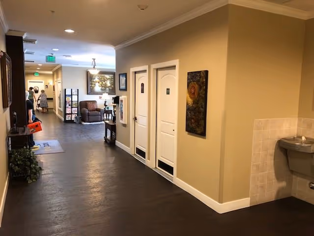 Well-lit interior hallway of a care facility with dark wood floors, two white doors and wall art on the right, a seating area at the end, and a wall-mounted water fountain.