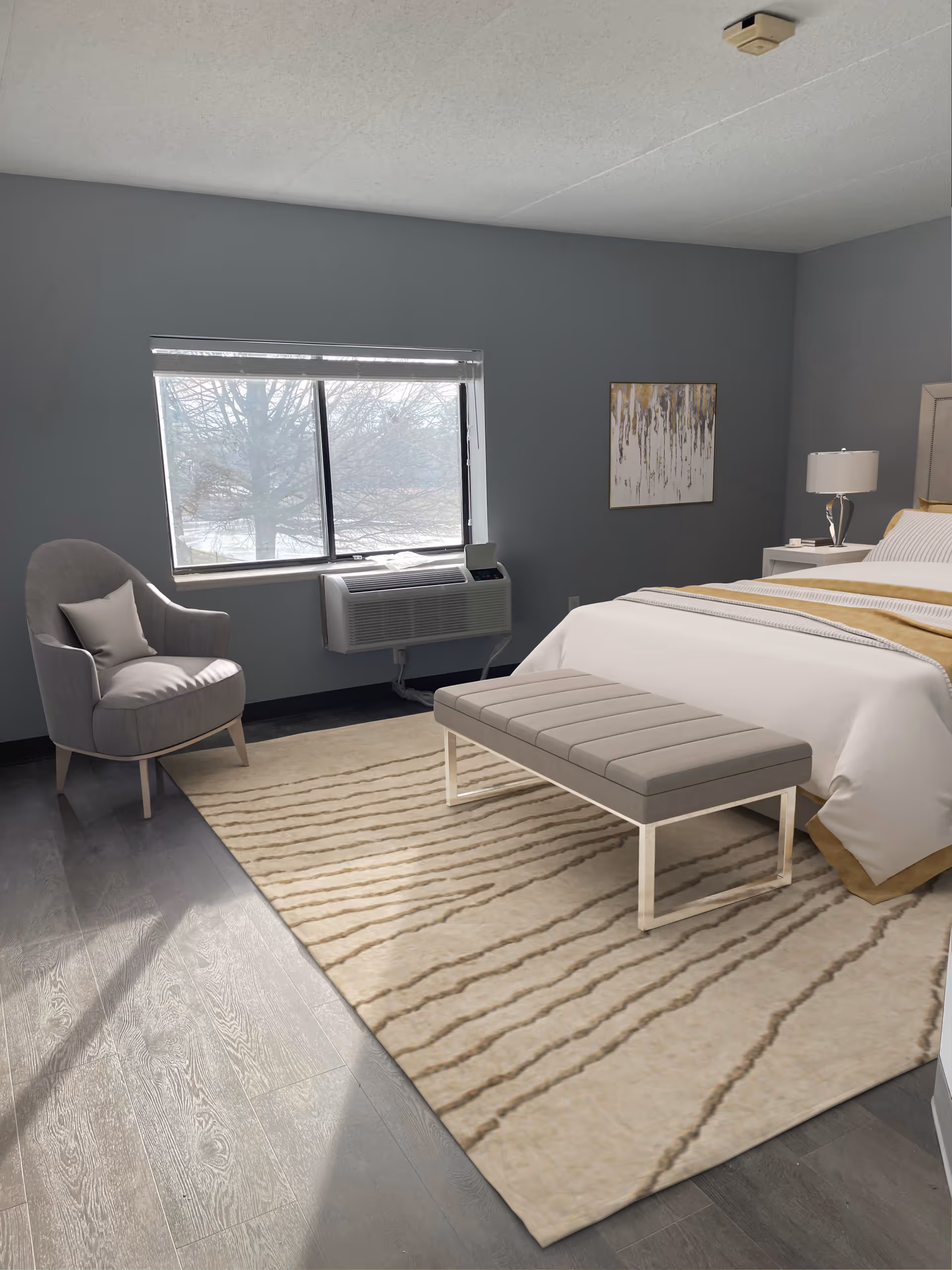 A bedroom with a large window showing a tree outside. The room has gray walls, a gray armchair with a small pillow, a beige and brown striped rug, a bench at the foot of the bed, and a bed with white and gold bedding. A nightstand with a lamp and a piece of abstract wall art are also visible.