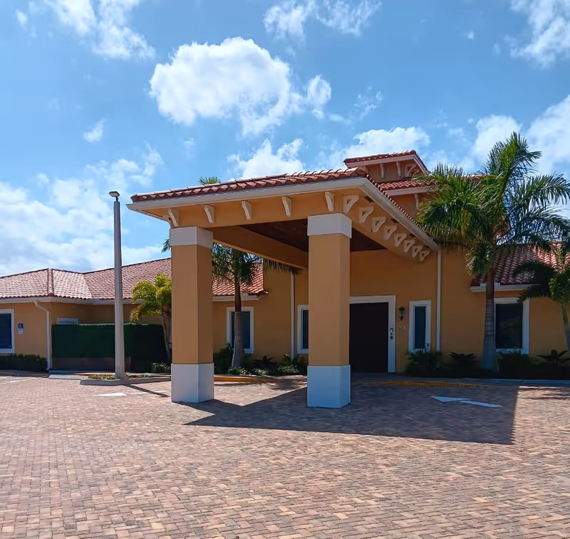 Entrance of a single-story Mediterranean-style building with a covered porte-cochere, palm trees, and a brick-paved driveway under a blue sky.