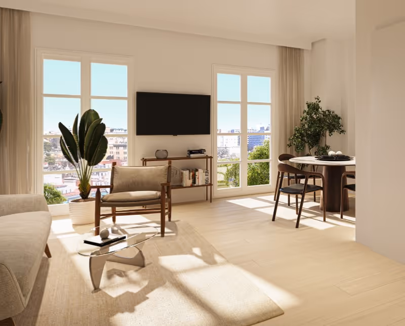 Bright and modern living room with large windows letting in natural light. The room features a beige sofa, a wooden armchair with cushions, a glass coffee table on a light rug, a wall-mounted flat-screen TV above a small wooden shelf with books and decor, and a round dining table with four chairs near a large potted plant.