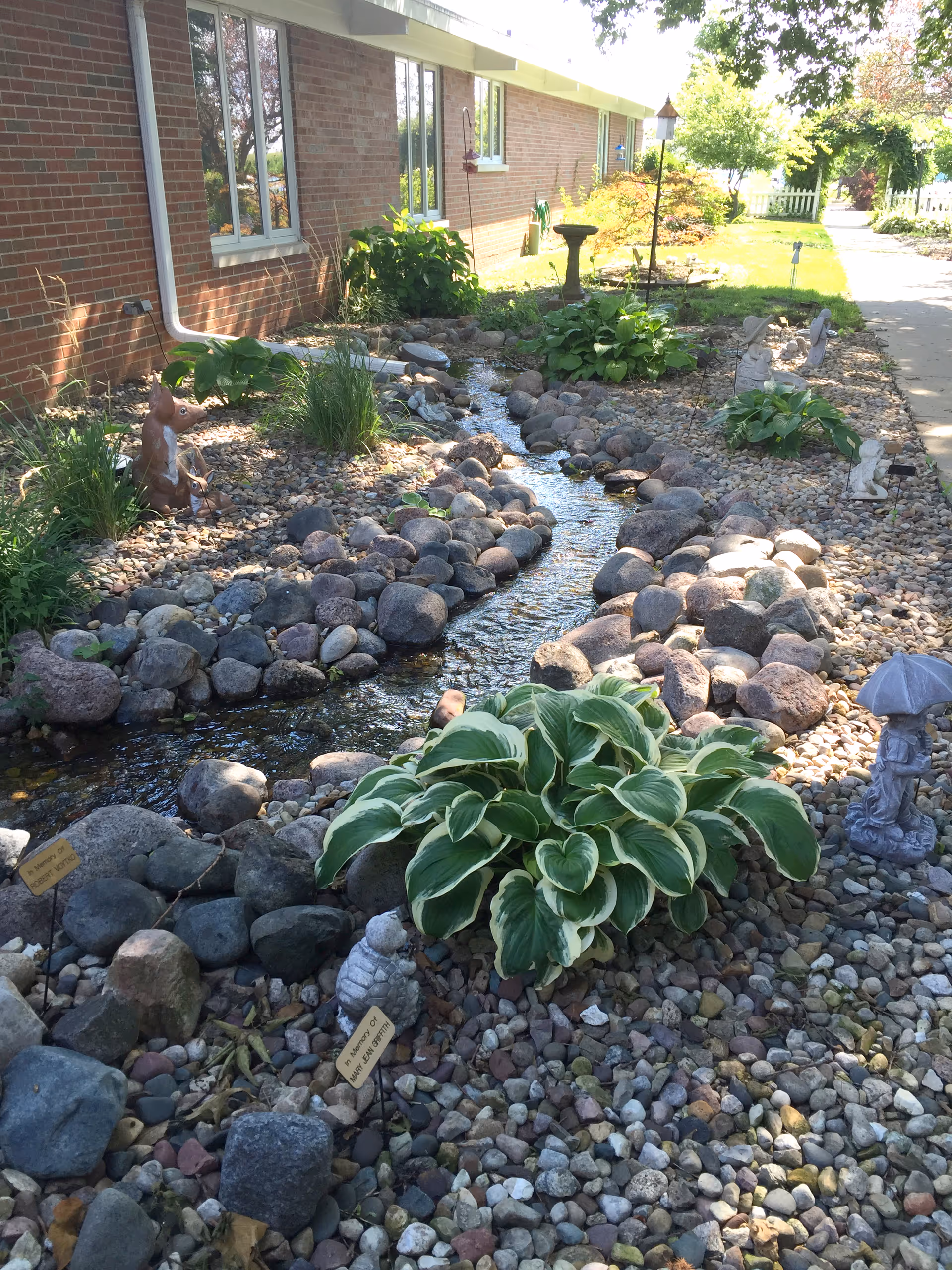 A landscaped garden area next to a brick building with a small artificial stream bordered by rocks and plants. There are decorative garden statues including a squirrel, a small figure holding an umbrella, and a birdbath. The garden is well-maintained with green plants and a pathway visible in the background.