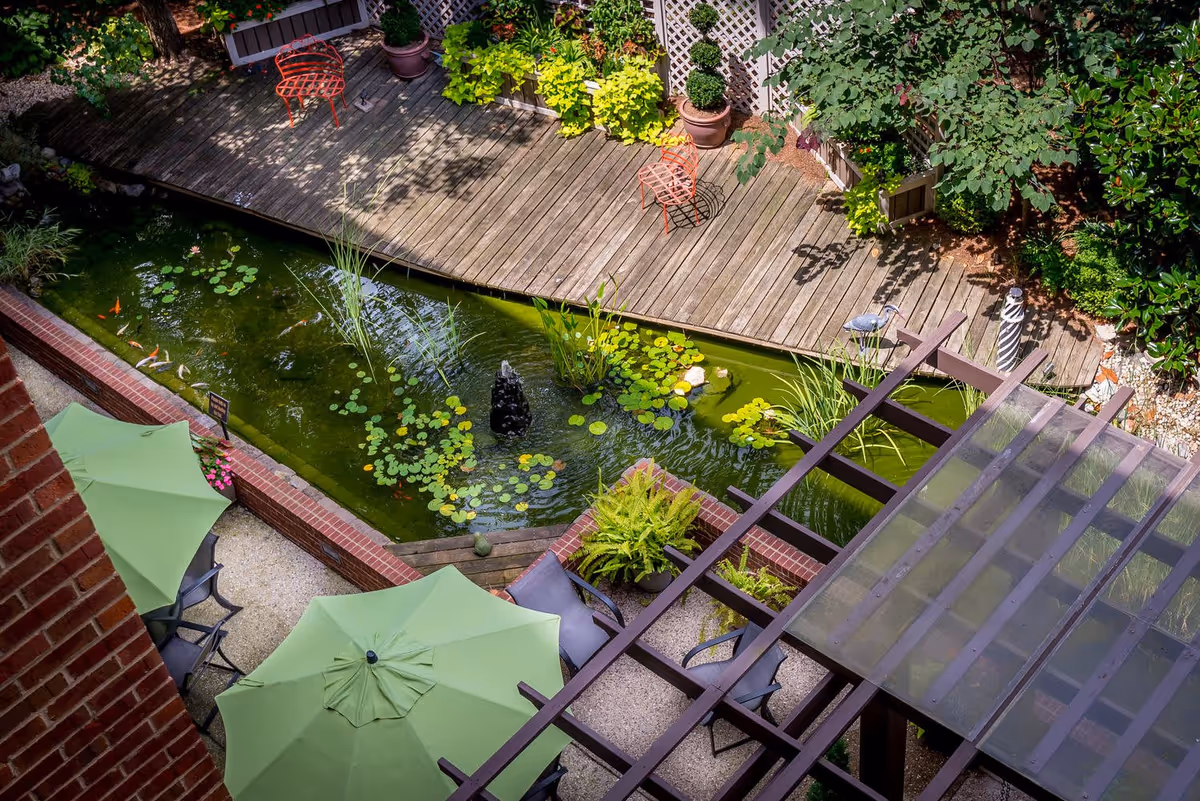 Overhead view of a serene outdoor garden area at Mayfair Village Retirement Community featuring a koi pond with lily pads and a small fountain, surrounded by a wooden deck with potted plants and two red chairs. Below the pond area, there are green patio umbrellas shading outdoor seating on a gravel surface, and a pergola with a translucent roof partially covering the seating area.