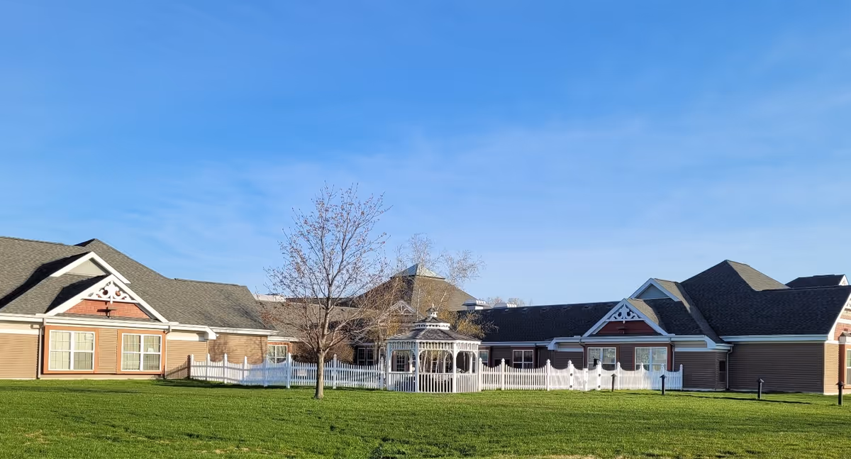 Exterior view of a single-story senior living facility building with a large green lawn in front, a white gazebo surrounded by a white picket fence, and a clear blue sky overhead.