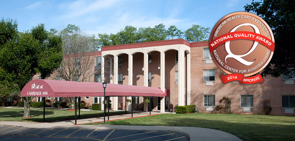 Exterior view of Carriage Inn of Steubenville, a three-story brick building with a covered entrance canopy in maroon color. The building is surrounded by green grass and trees under a clear blue sky.