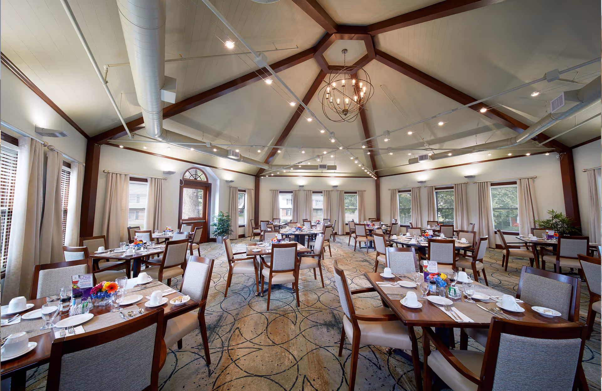 A spacious dining room with multiple wooden tables and cushioned chairs arranged neatly. Each table is set with white plates, cups, silverware, and glasses. The room features large windows with beige curtains, a high vaulted ceiling with exposed beams, modern light fixtures, and a patterned carpeted floor.