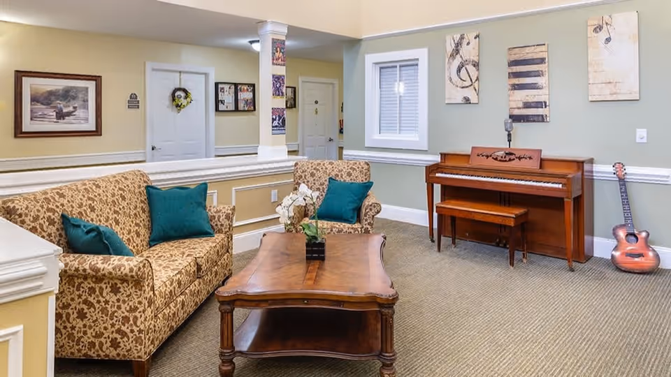 A cozy living room area with a patterned sofa and armchair, both adorned with teal cushions, a wooden coffee table with a small plant, a wooden piano with a bench, and a guitar resting against the wall. The walls are decorated with musical-themed artwork and a window with white blinds.
