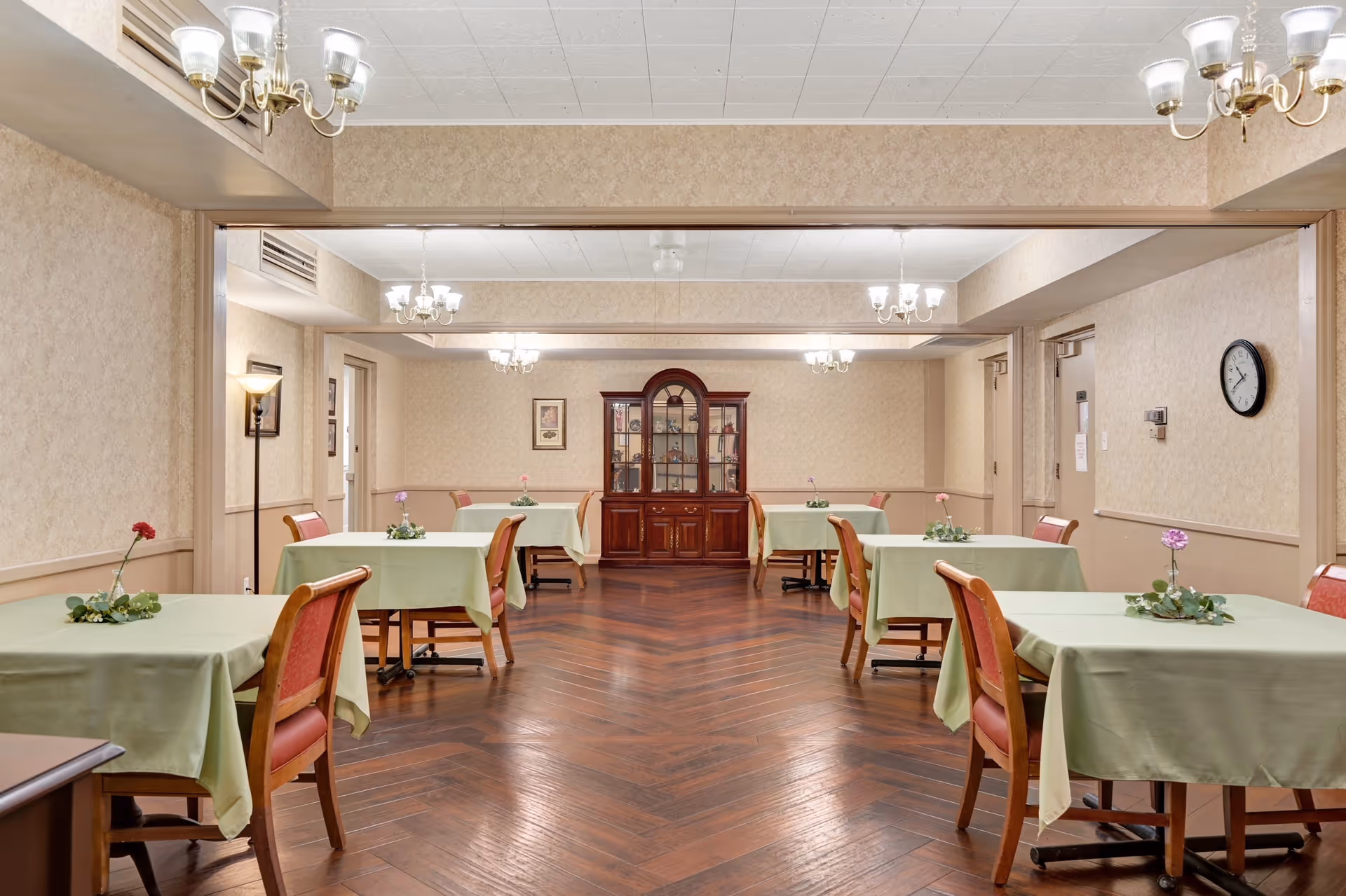 A dining room in a senior living facility with several tables covered in light green tablecloths, each adorned with a small flower arrangement. The room has wooden flooring with a herringbone pattern, beige wallpapered walls, and multiple chandeliers providing lighting. A wooden china cabinet is positioned against the far wall, and a clock is mounted on the right wall.