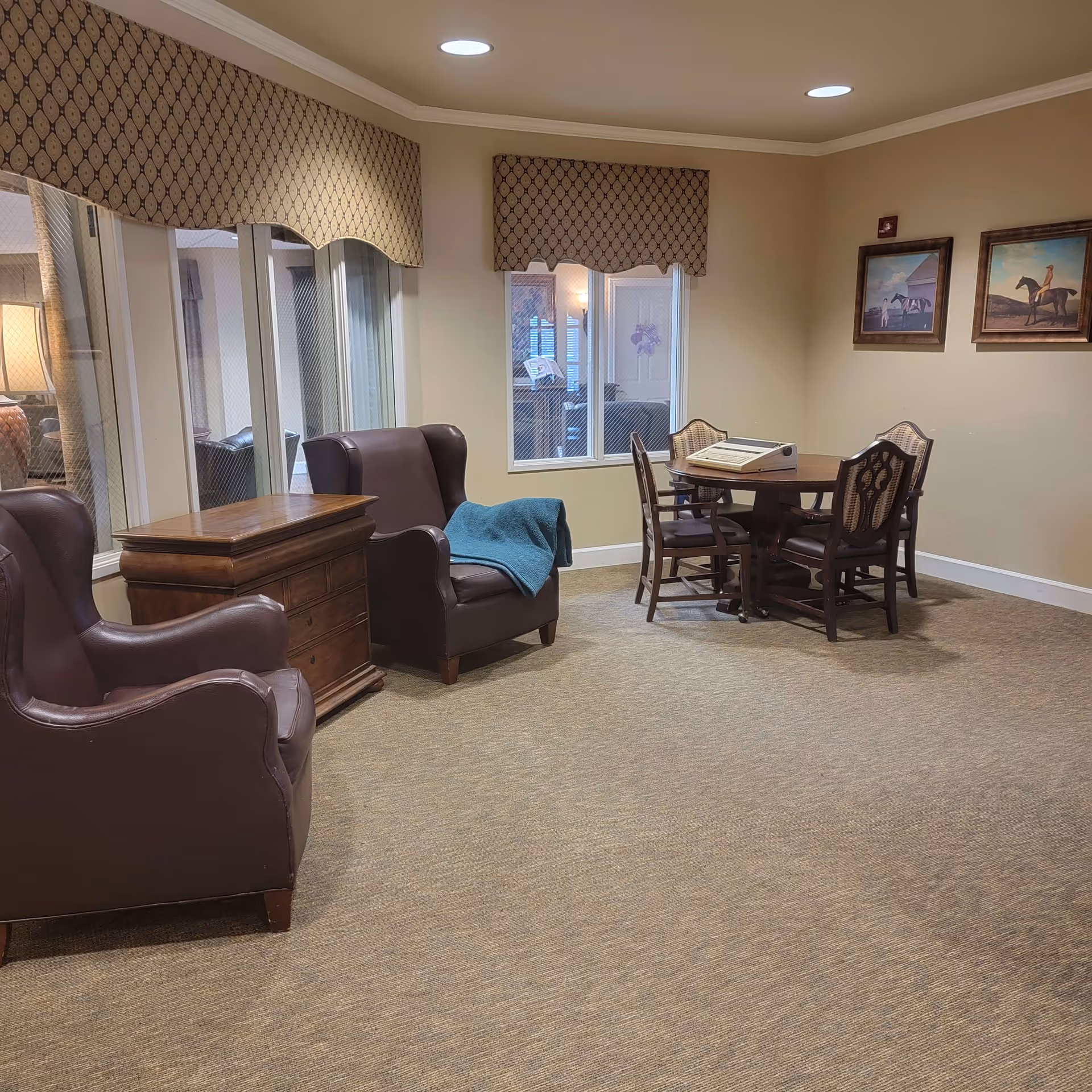 A cozy sitting area in a senior living facility with two brown leather armchairs, a wooden side table, and a round wooden table with four chairs. The room has beige walls, carpeted floor, patterned valances on the windows, and two framed paintings of horses on the wall.