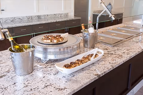 Granite buffet counter displaying platters of hors d'oeuvres and wine bottles chilling in metal buckets under a glass sneeze guard.