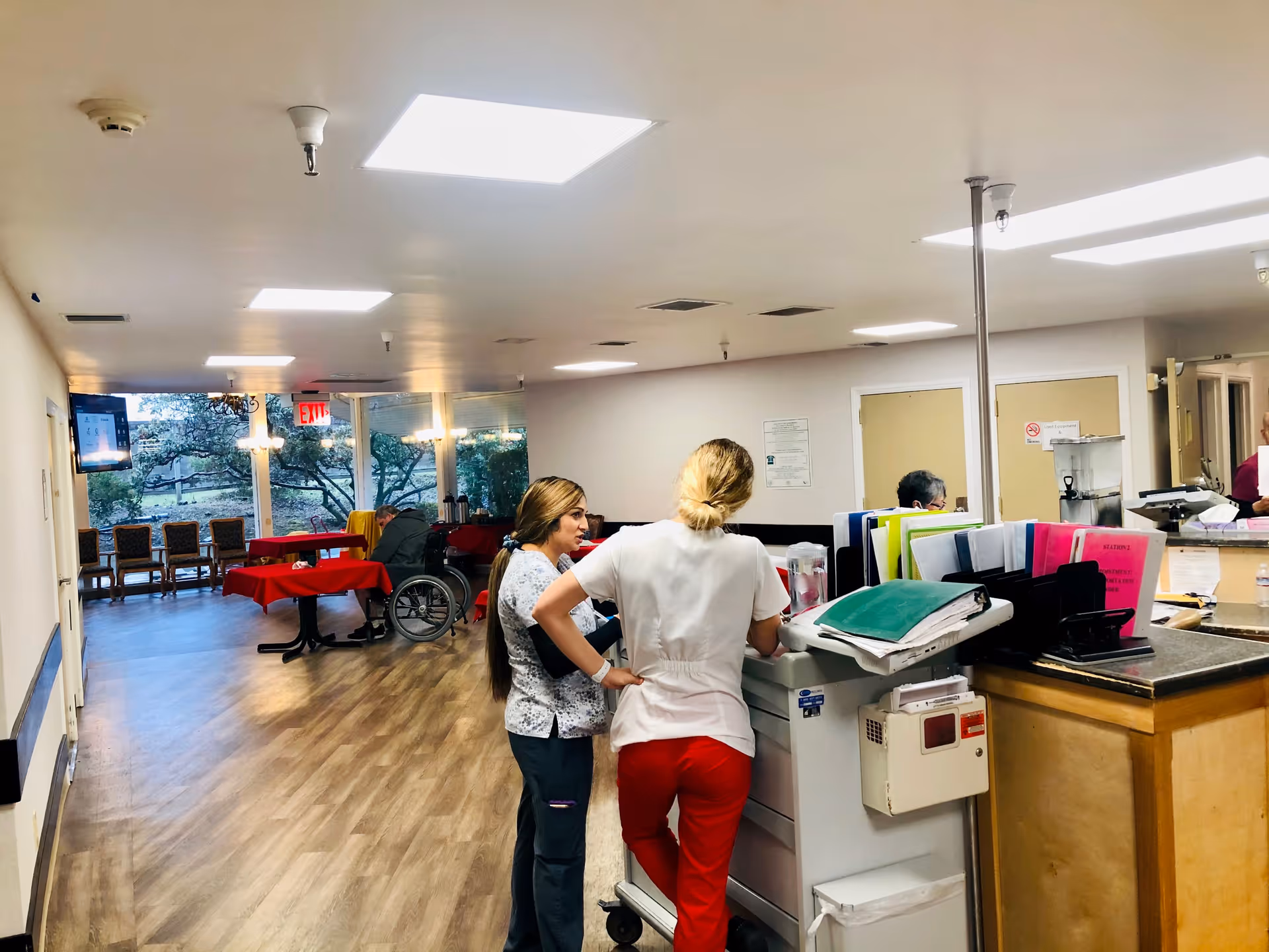 Interior view of a transitional care center showing two healthcare workers standing and talking near a workstation with files and medical equipment. In the background, there are tables covered with red tablecloths, chairs, a person in a wheelchair, and large windows showing trees outside.