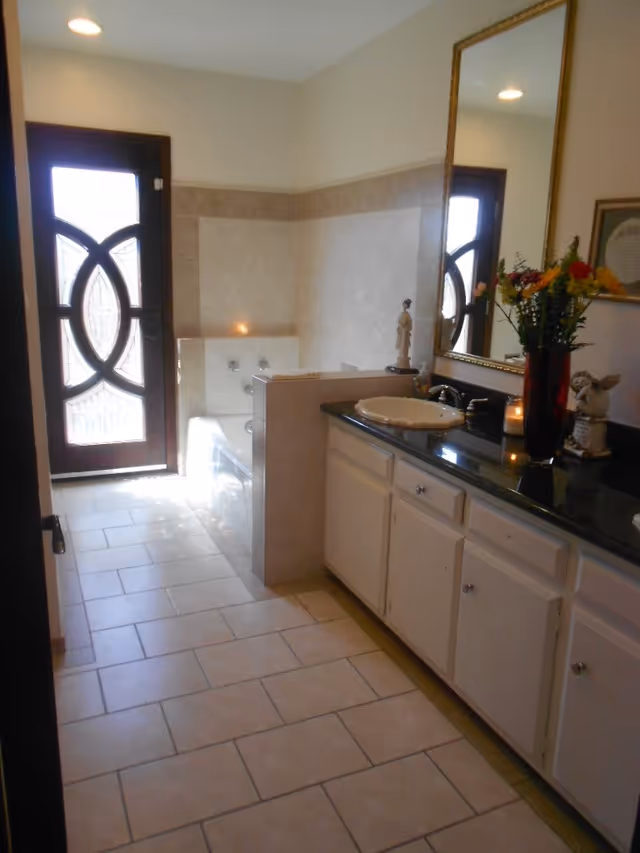 A bathroom with beige tiled floor and walls, featuring a bathtub with jets, a black countertop with a white sink, a large mirror above the sink, decorative items including a vase with flowers, candles, and small statues. A door with decorative glass panels is visible at the end of the room.