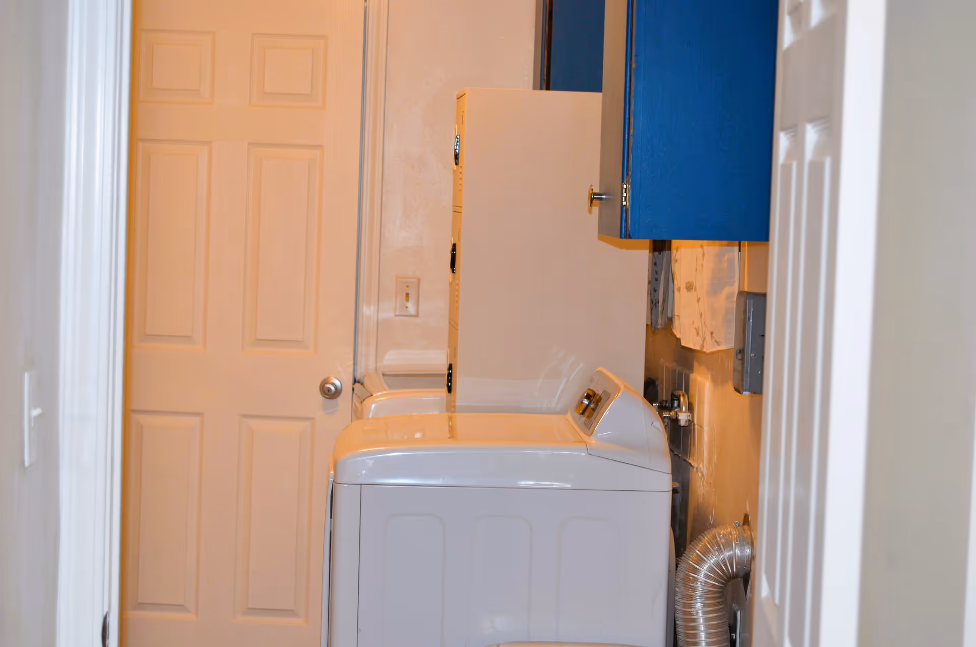 A laundry room with a white washing machine and dryer side by side. Above the machines are blue wall-mounted cabinets. There is a closed beige door at the end of the room and a vent hose connected to the dryer.