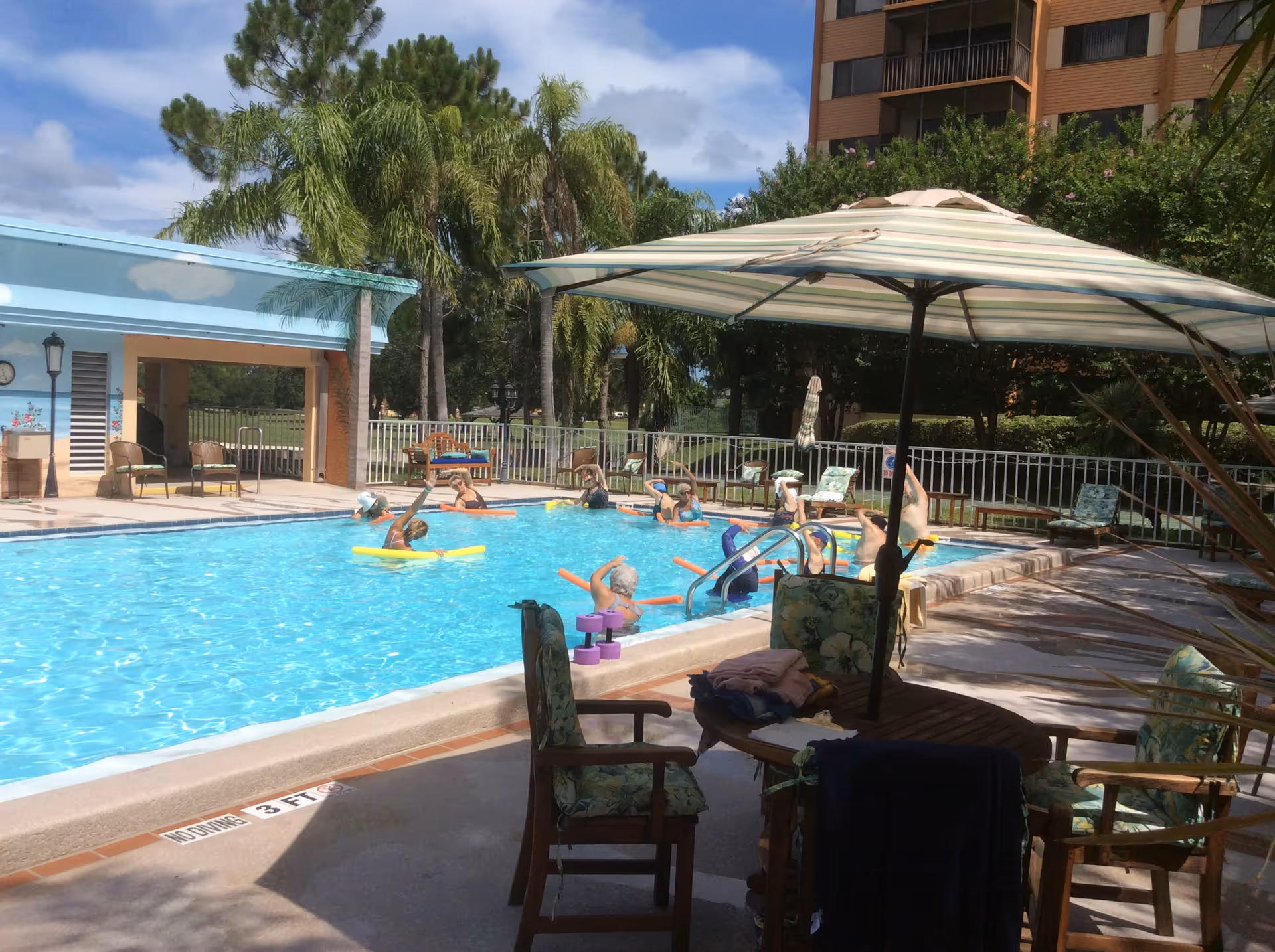 Outdoor swimming pool at a senior living facility with residents doing water aerobics under umbrellas and palm trees.