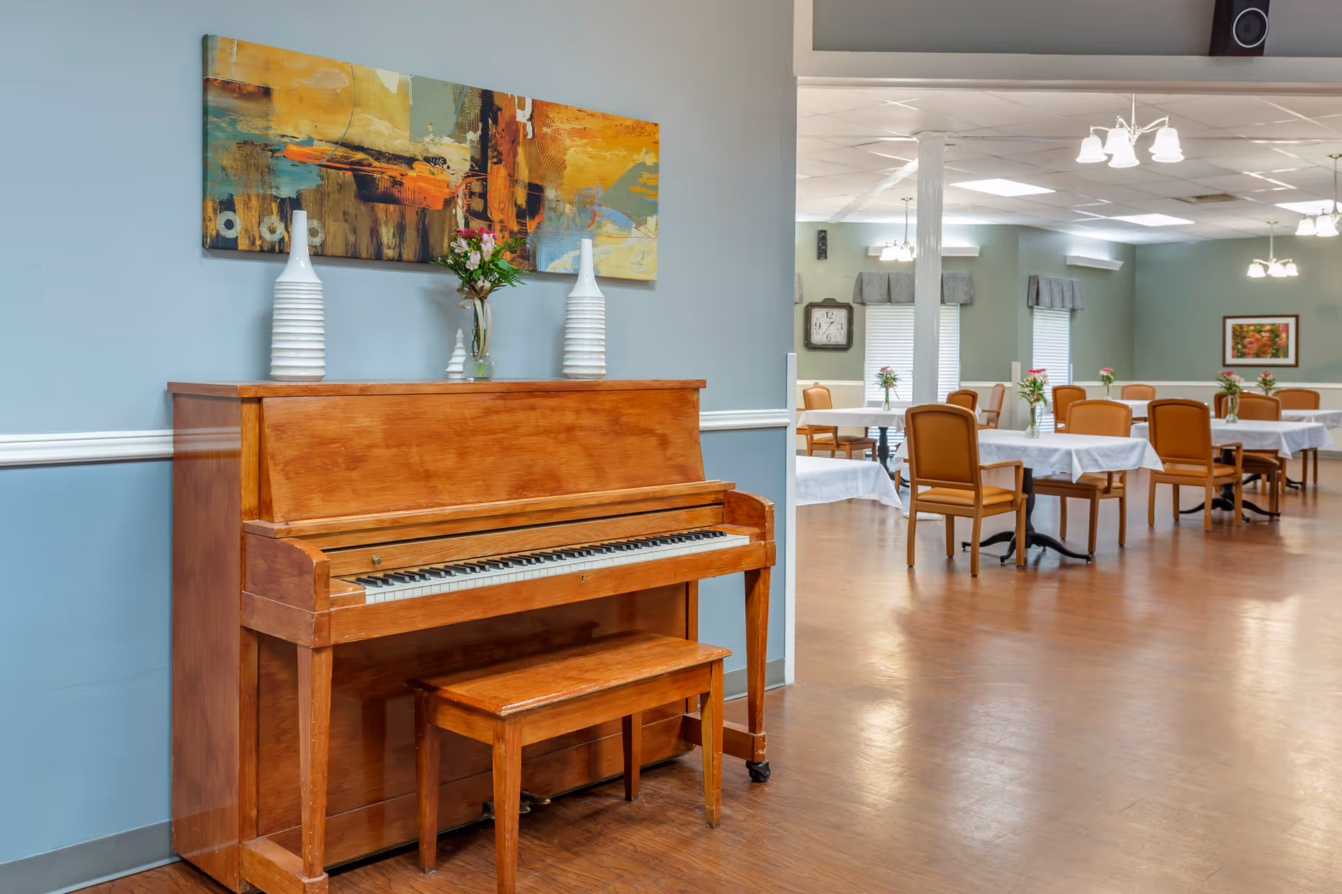 A wooden upright piano with a matching bench is placed against a light blue wall. On top of the piano are two white decorative vases and a small glass vase with flowers. Above the piano hangs a colorful abstract painting. In the background, there is a dining area with tables covered in white tablecloths, each adorned with a small vase of flowers. The dining chairs are wooden with cushioned seats, and the room has large windows with valances and ceiling lights.