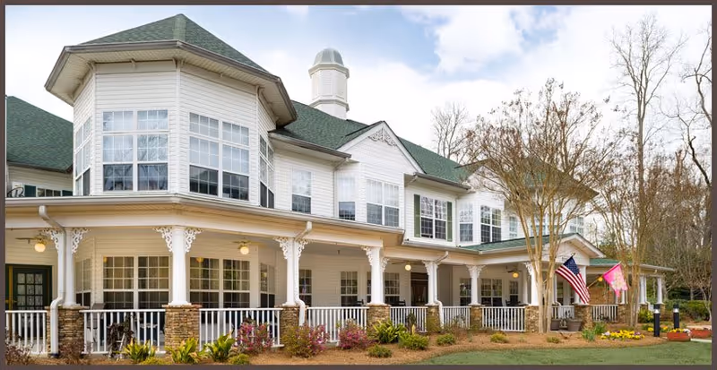 Exterior view of a large two-story senior living facility building with white siding, green roof, and a wraparound porch with white railings and decorative columns. There are American and other flags displayed near the entrance, landscaping with bushes and flowers, and trees without leaves in the background under a partly cloudy sky.