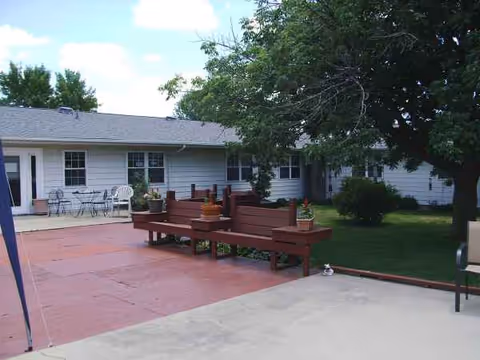 Single-story light-gray building with red wooden benches on a shaded outdoor patio.